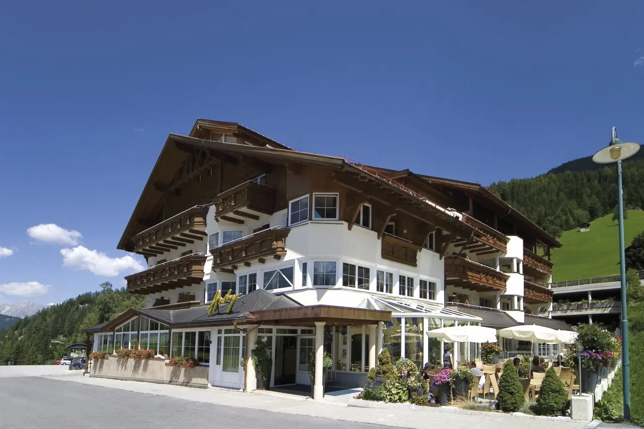Aussenansicht des Hotel Andy im alpinen Stil mit sonniger Terrasse, Blumen und grünen Bergen im Hintergrund.