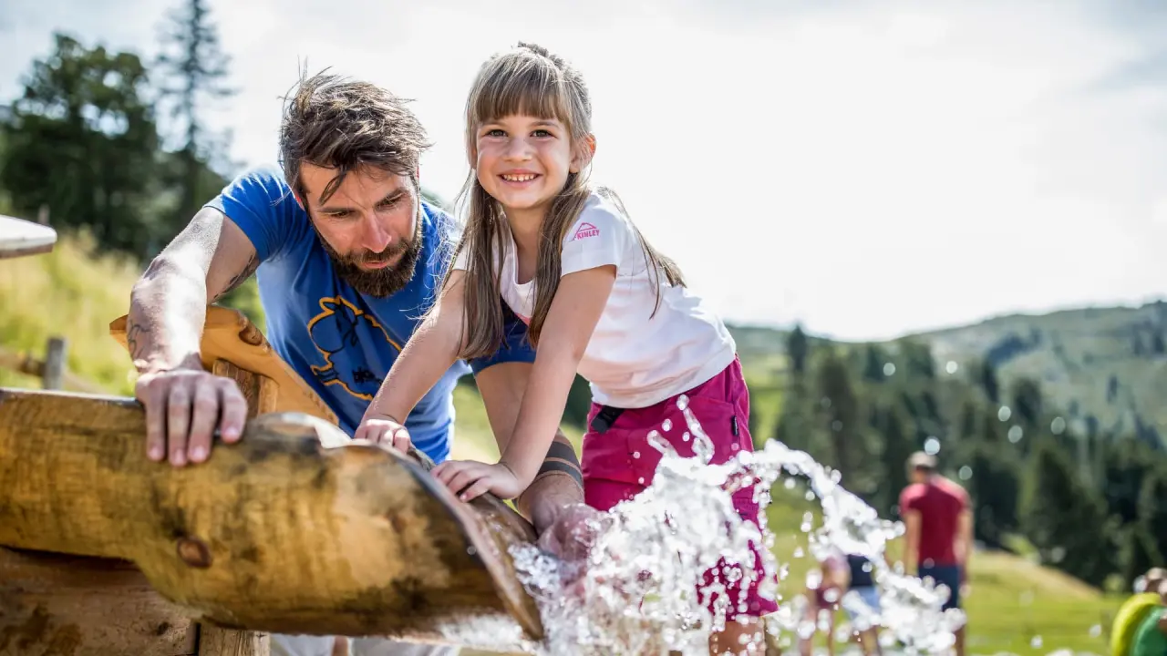 Lachendes Mädchen und Vater spielen mit Wasser in einem Holzbrunnen im Garten des Hotel Andy.