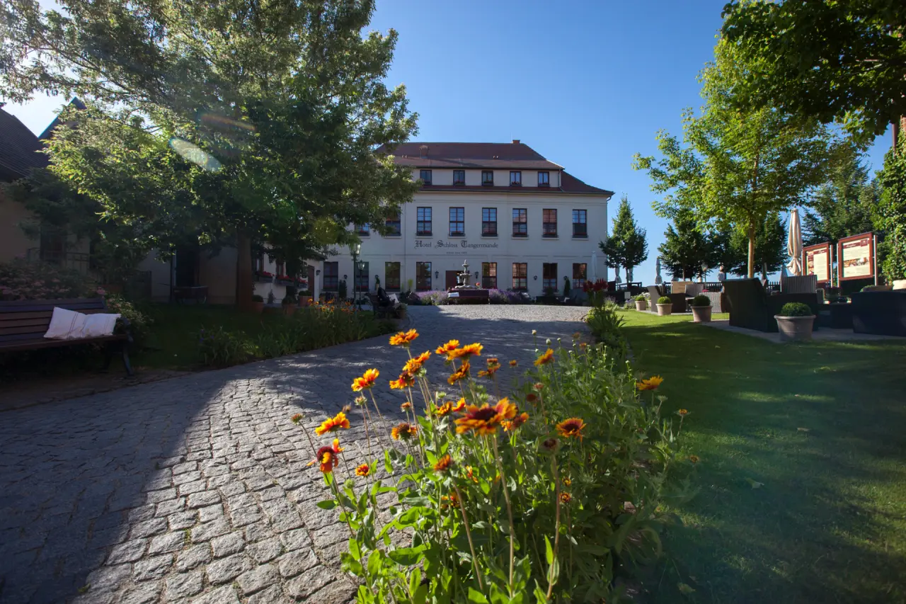 Aussenansicht des Hotel Schloss Tangermünde mit Kopfsteinpflasterweg, blühenden Blumen und Gartenbereich unter blauem Himmel.