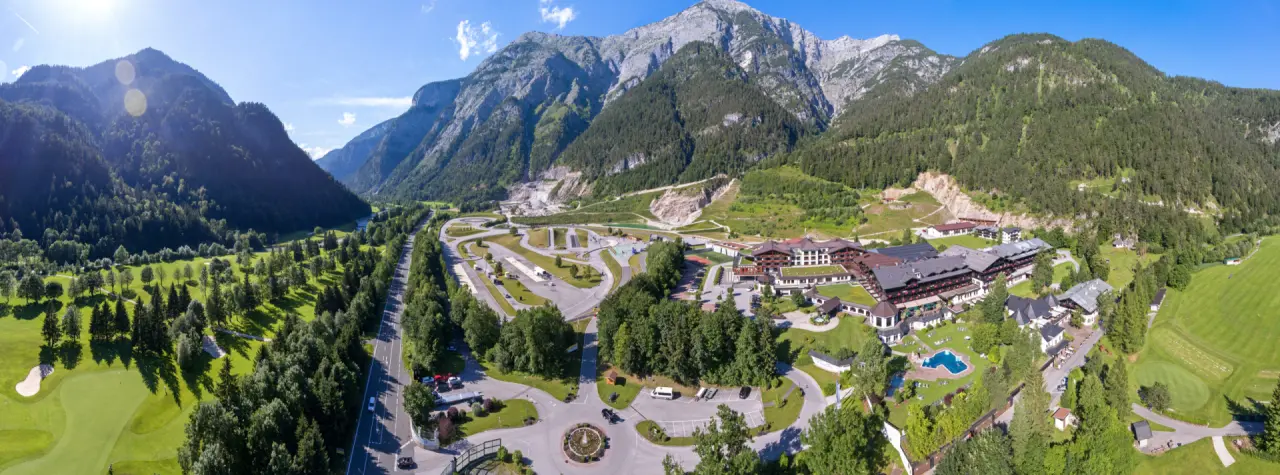 Luftaufnahme des Hotel Gut Brandlhof mit weitläufiger Anlage, Outdoor-Pool, Golfplatz und majestätischer Bergkulisse.