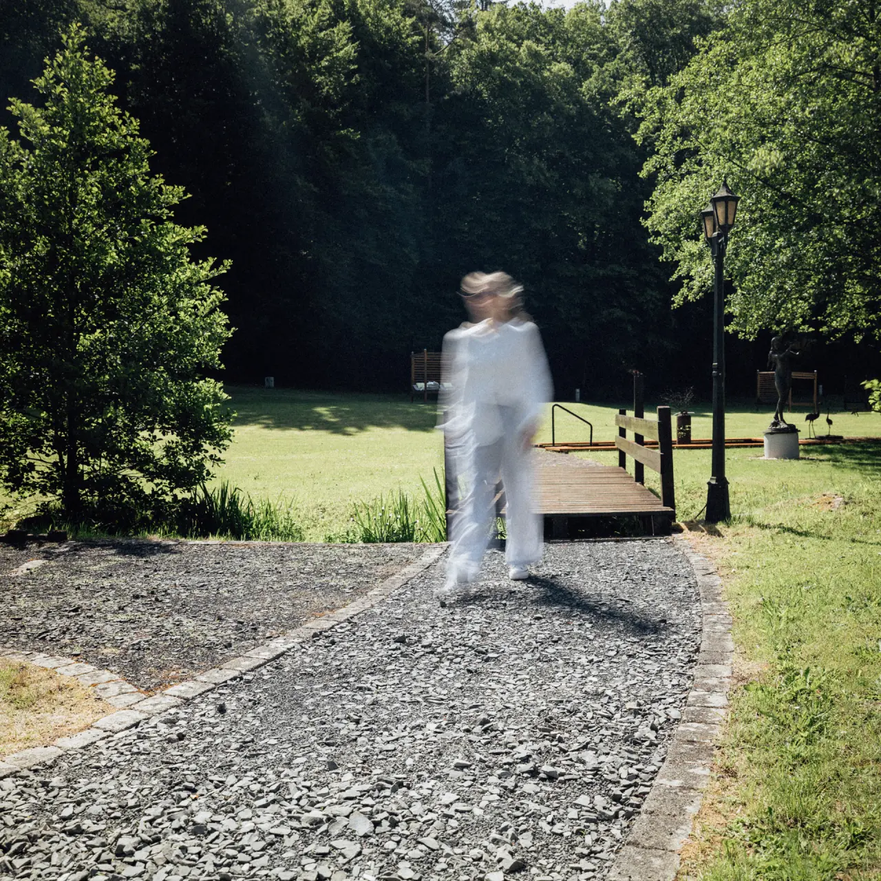 Verschwommene Person auf Kiesweg im weitläufigen Garten des Hotel Im Krummbachtal mit Bäumen und Holzsteg.