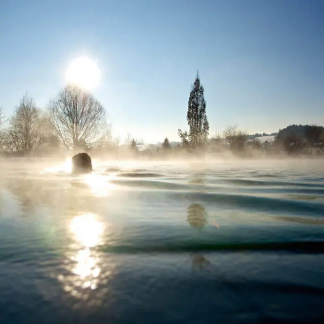 Person schwimmt im dampfenden Aussenpool des Tannenhof Sport & SPA bei Sonnenaufgang im Winter.