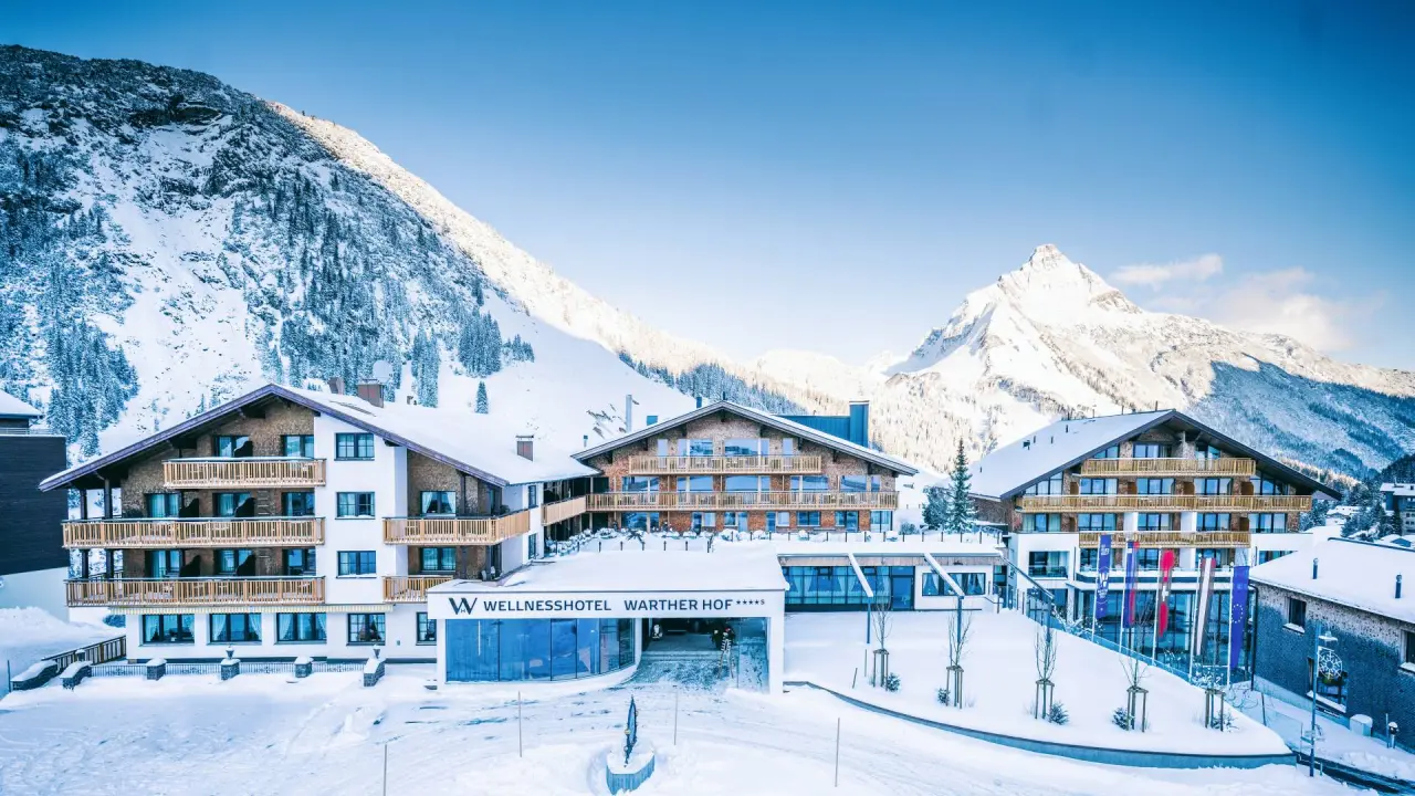 Majestätische Aussenansicht des Wellnesshotel Warther Hof im Winter, umgeben von schneebedeckten Alpen unter blauem Himmel.