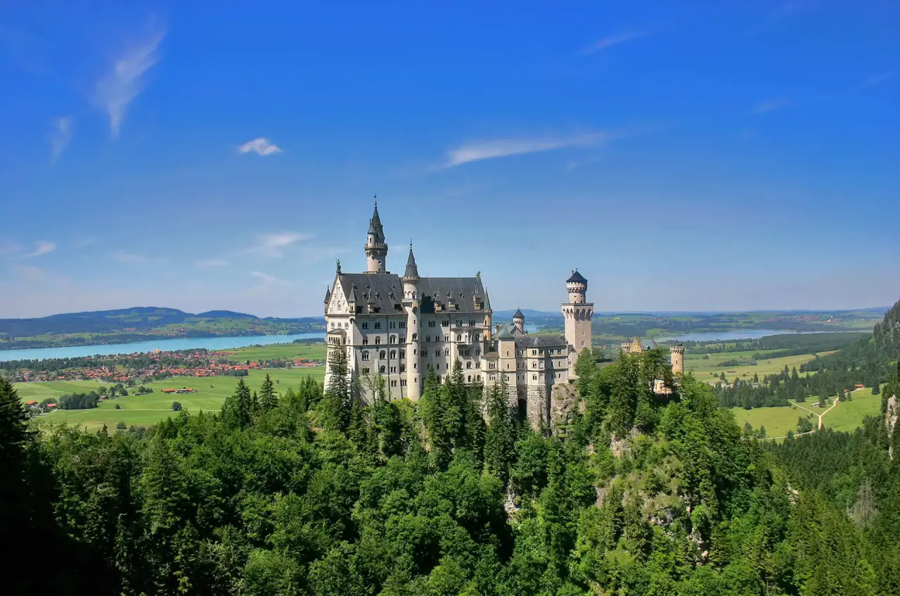 Schloss Neuschwanstein in den bayerischen Alpen mit Seen und grünen Wäldern unter blauem Himmel.