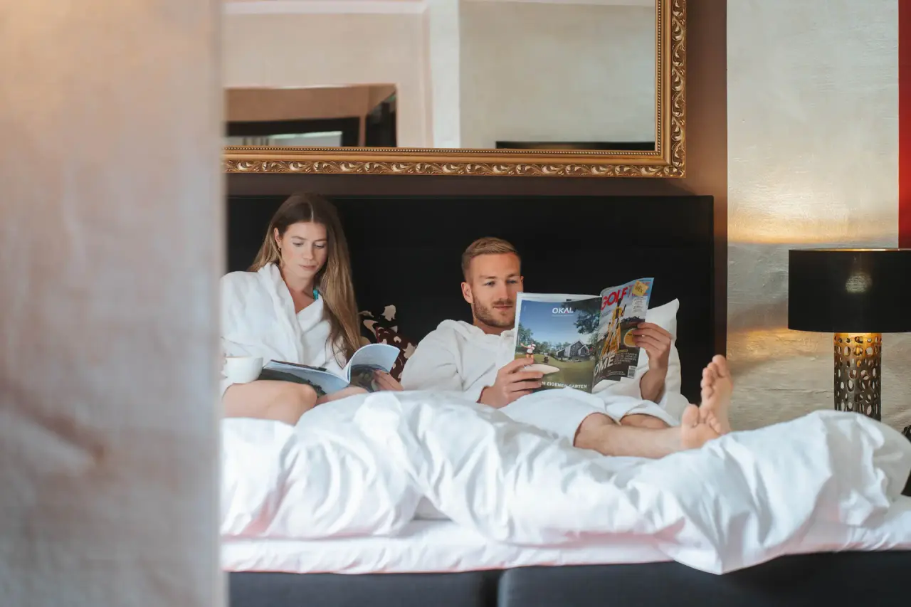 Paar in Bademänteln entspannt lesend im komfortablen Hotelzimmer des Golf- & Alpin Wellness Resort Hotel Ludwig Royal.