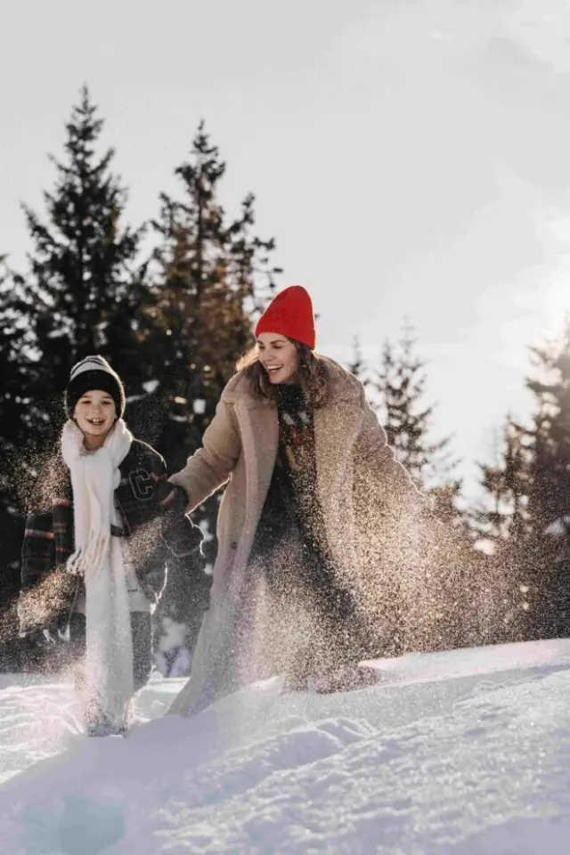 Lachende Mutter und Kind spielen im tiefen Schnee vor winterlicher Waldkulisse, Hotel Oberforsthof.