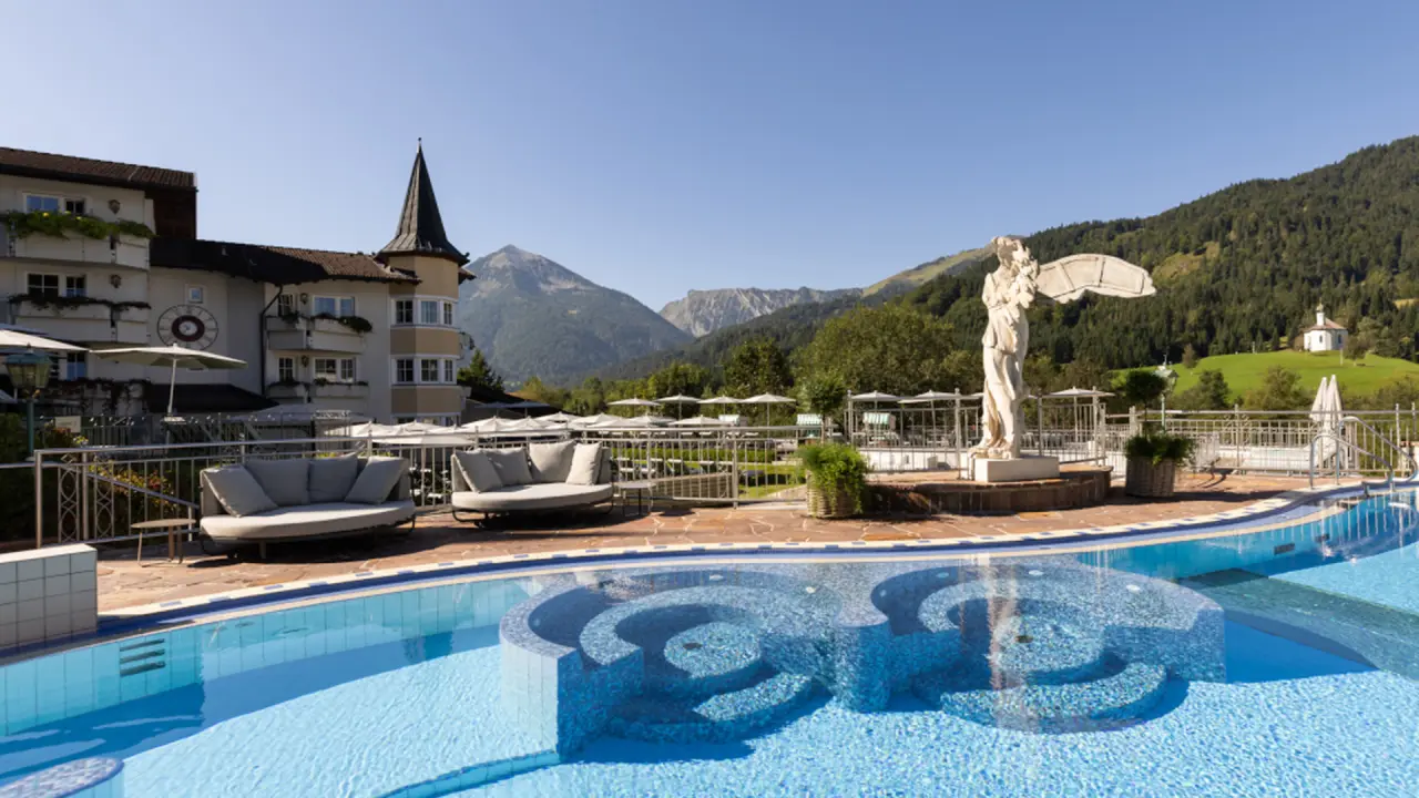 Aussenpool des Posthotel Achenkirch mit eleganten Liegen, umgeben von majestätischen Bergen und grünen Wäldern unter blauem Himmel.