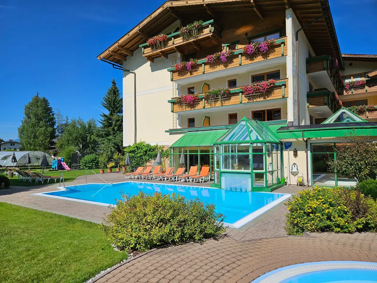 Aussenansicht des Vitalhotel Berghof mit sonnigem Aussenpool, Liegen und blühenden Balkonen vor blauem Himmel.