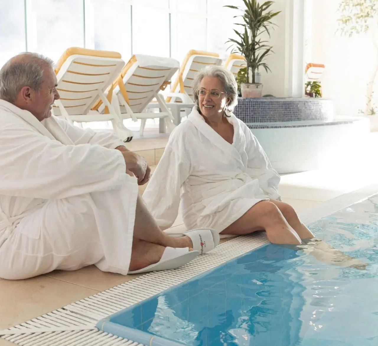 Entspanntes Paar in Bademänteln am Pool im Vitalhotel Berghof. Gäste genießen den Wellnessbereich mit Sonnenliegen.