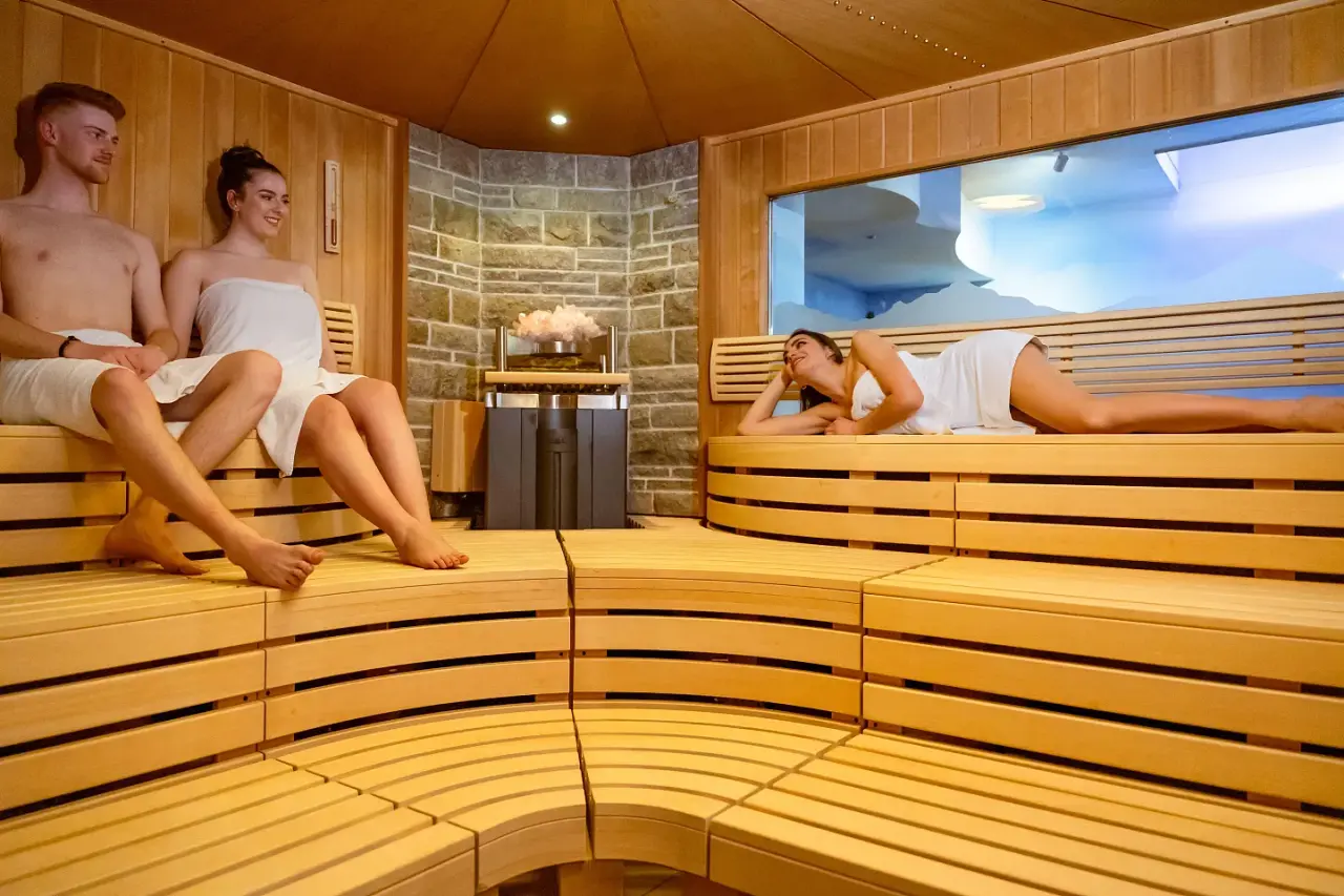 Moderne finnische Sauna mit Steinwand und entspannten Gästen im Vitalhotel Berghof Spa-Bereich.