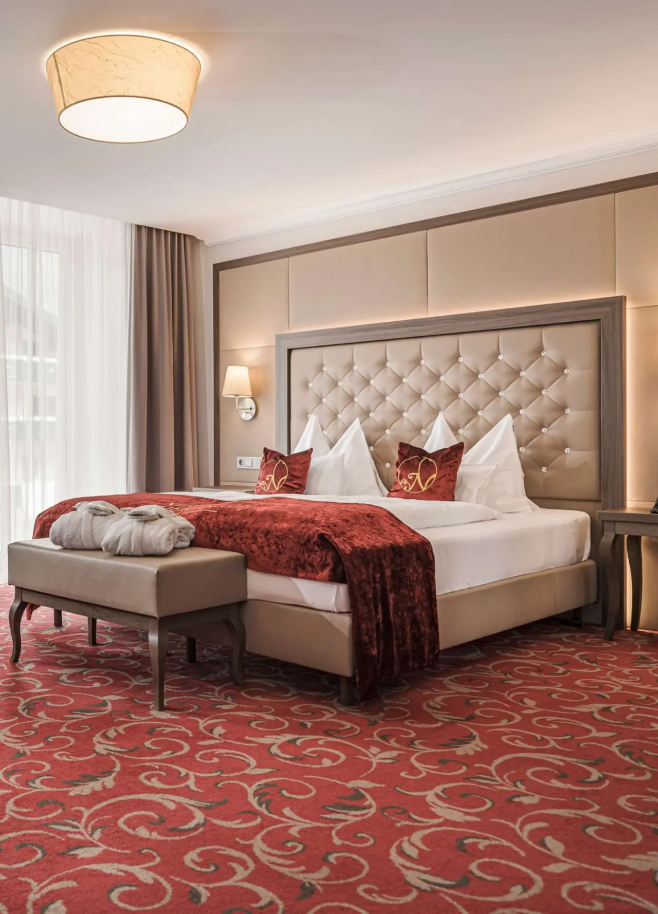Elegantes Doppelzimmer im Hotel Norica mit großem Bett, roten Akzenten und Bademänteln auf der Sitzbank.