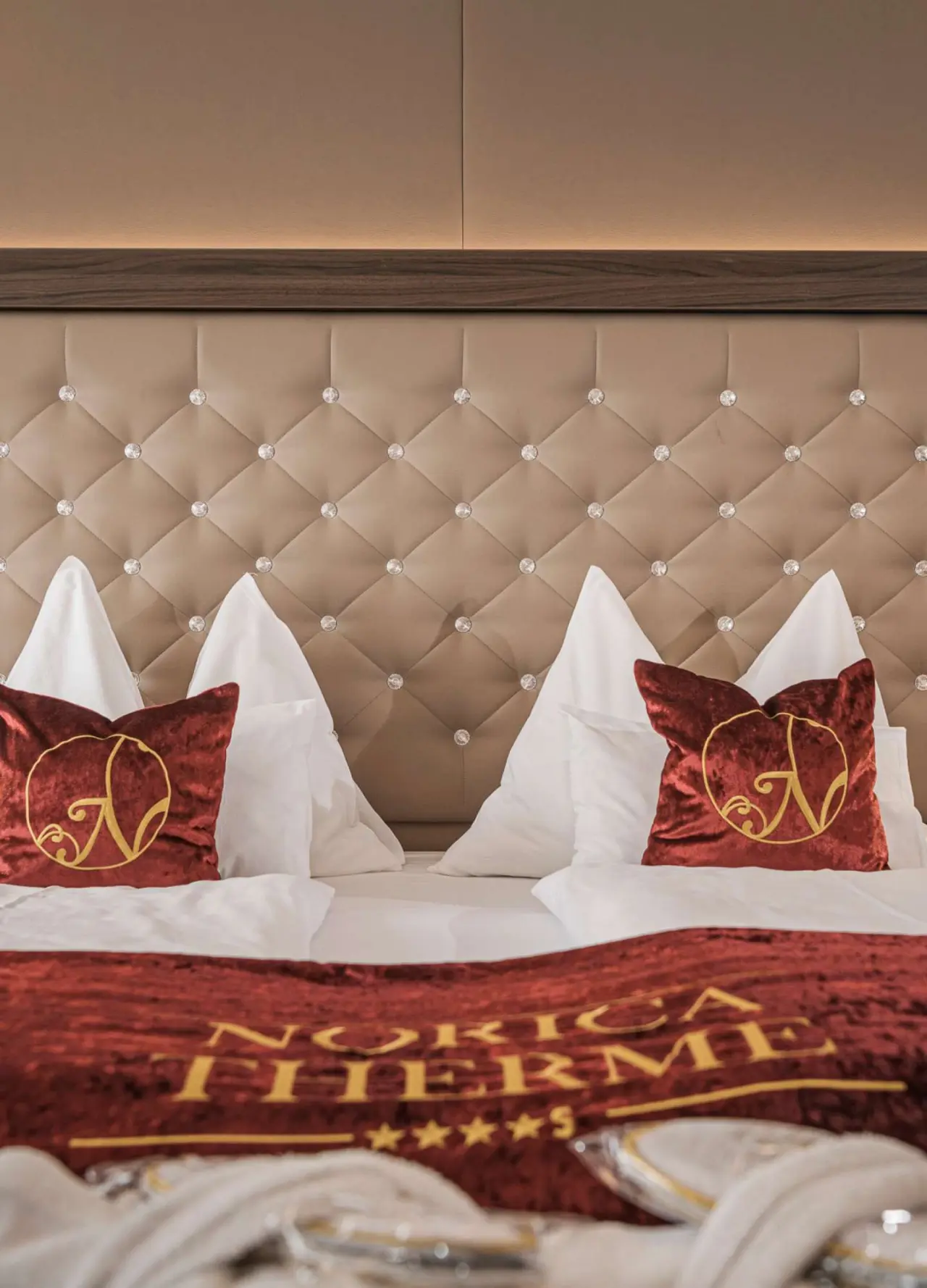 Luxuriöses Doppelbett mit gepolstertem Kopfteil und roten Kissen mit Hotel Norica Logo im Hotel Norica Therme.