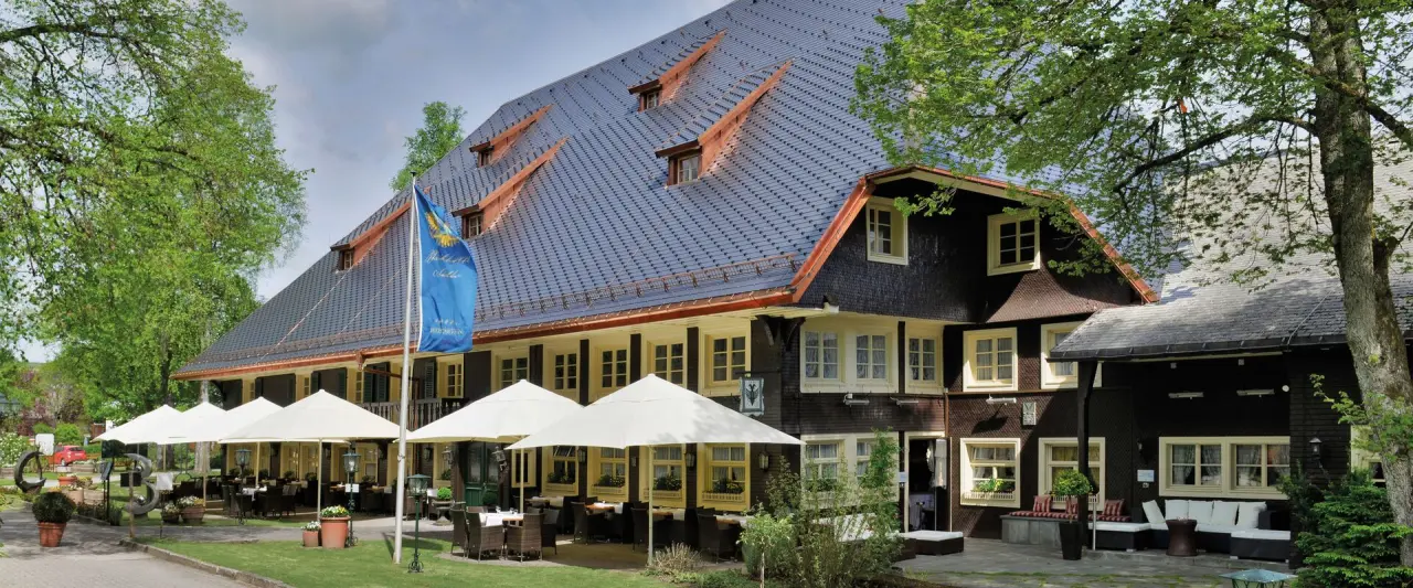 Aussenansicht des Parkhotel Adler mit traditioneller Architektur, blauer Flagge, grosser Terrasse mit Schirmen und üppigem Grün.