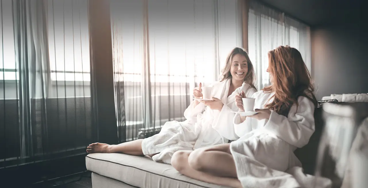 Zwei Frauen in Bademänteln entspannen lachend mit Kaffee in einer gemütlichen Lounge im TRIFORÊT alpin.resort.