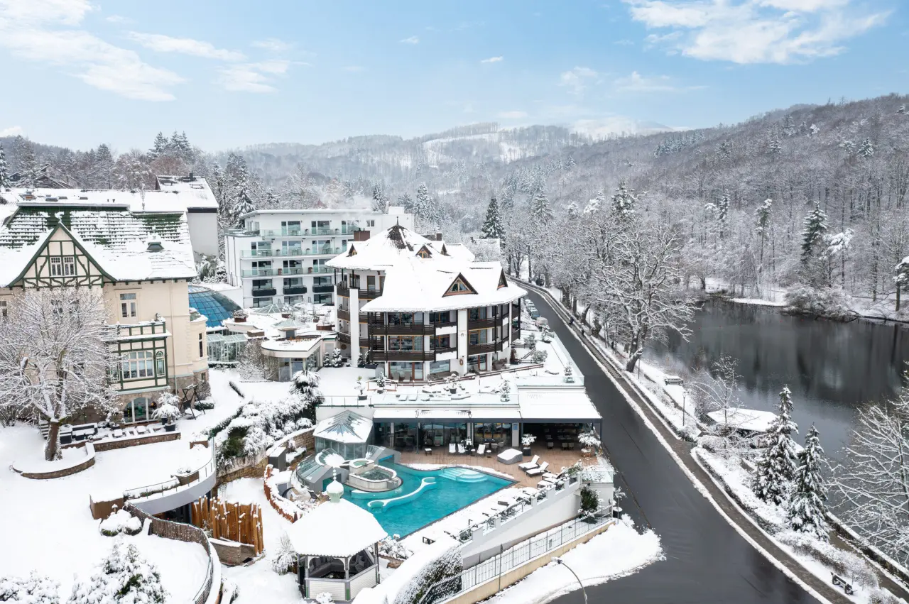 Winterlandschaft des Hotel Romantischer Winkel - RoLigio® & Wellness Resort mit verschneiten Gebäuden, blauem Aussenpool und See.