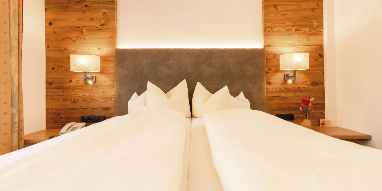 Gemütliches Doppelbett mit Holzwand und Leselampen im Zimmer des Aktiv- & Vitalhotel Bergcristall.
