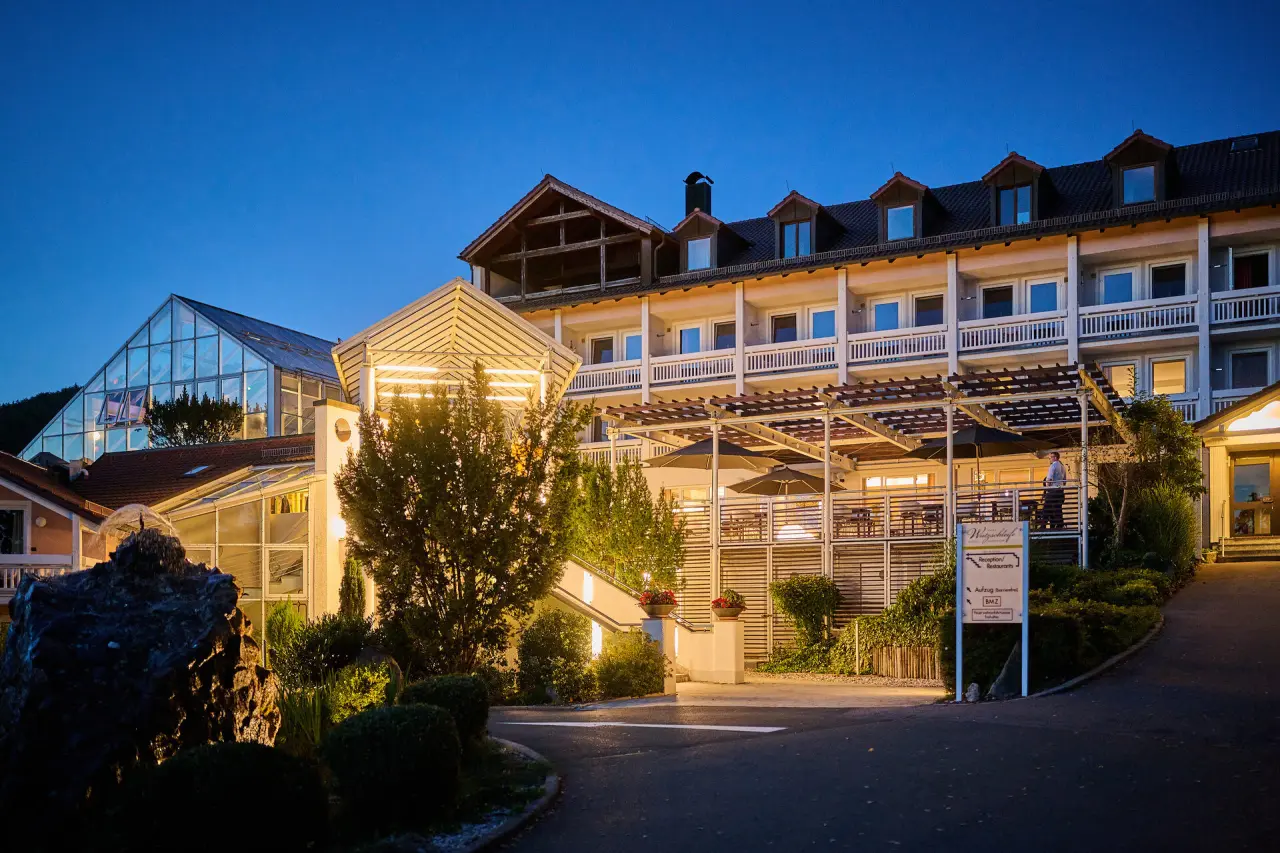 Abendansicht des Hotel Wutzschleife mit beleuchteter Fassade und großem Glasanbau unter blauem Himmel.