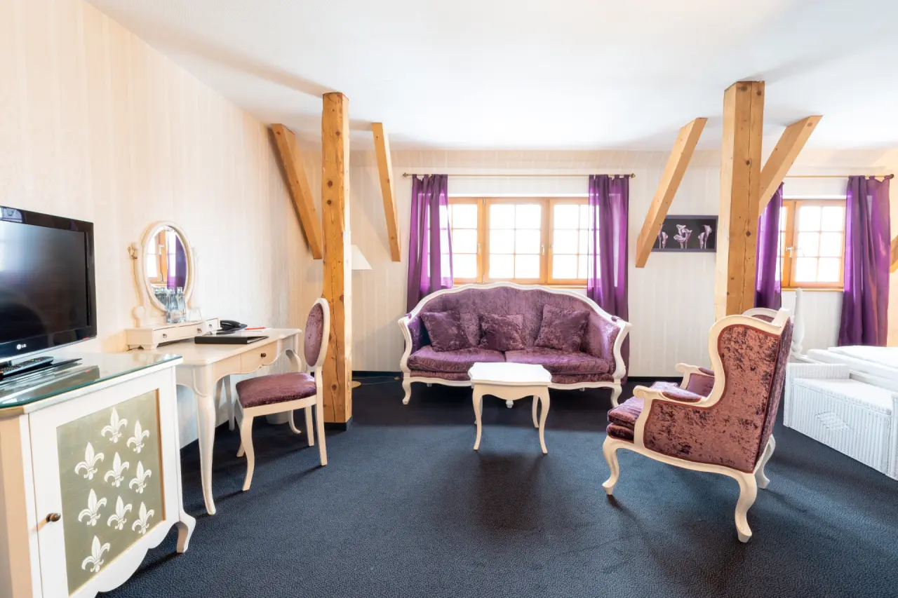 Geräumiges Hotelzimmer im Hotel & Spa Wasserschloss Westerburg mit lila Samtmöbeln, Holzbalken und Schreibtisch.