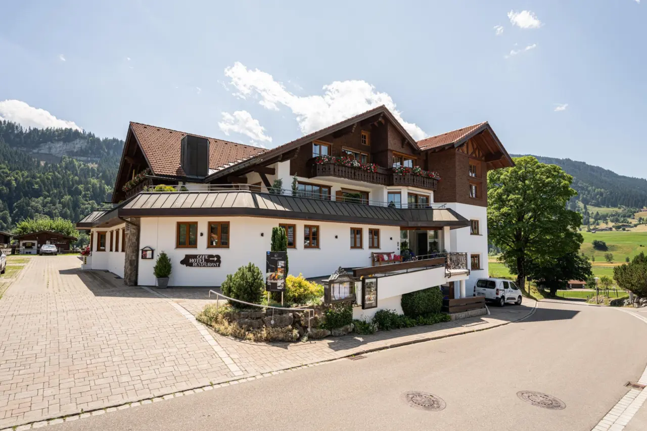 Aussenansicht des Hotel Berwanger Hof im alpinen Stil mit blühenden Balkonen und Bergkulisse bei Sonnenschein.