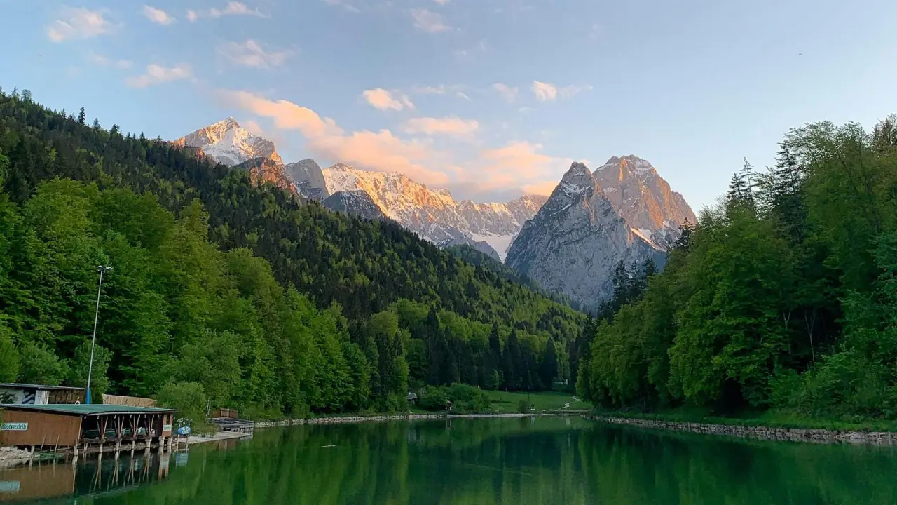Malerischer Riessersee mit spiegelnder Wasseroberfläche, umgeben von grünen Wäldern und majestätischen Alpen im Sonnenlicht.