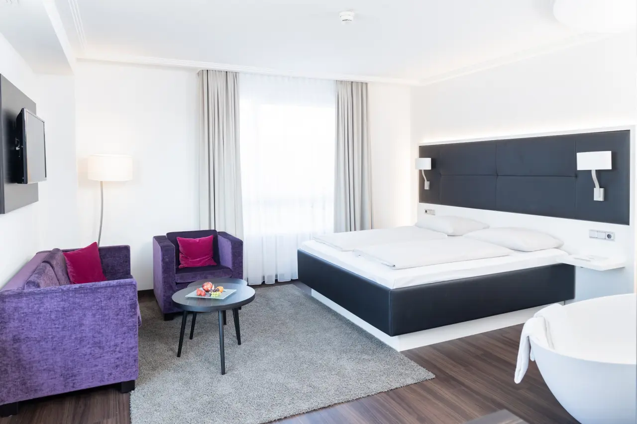 Modernes Hotelzimmer im Hotel Esperanto mit Doppelbett, lila Sitzbereich und freistehender Badewanne für entspannte Aufenthalte.