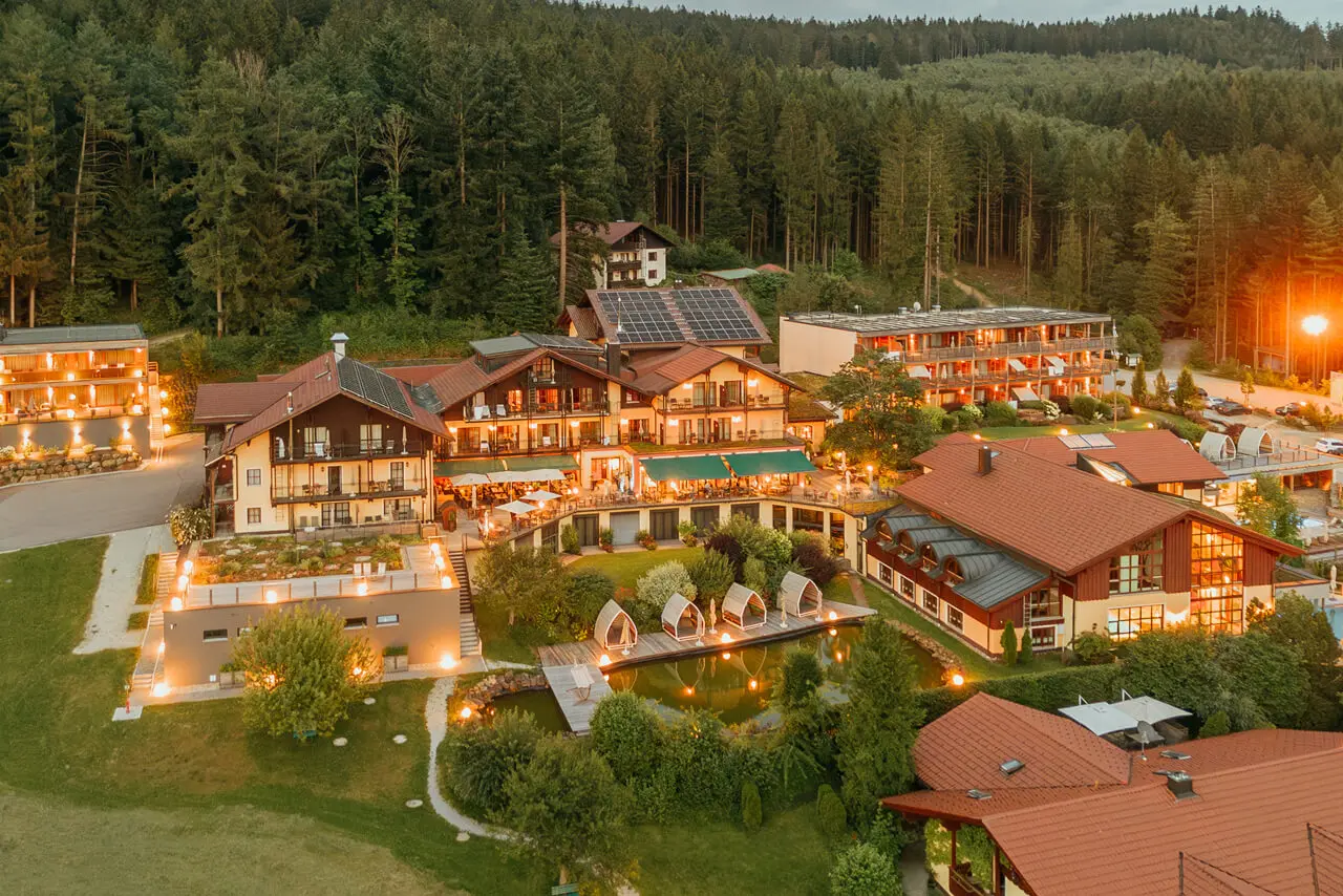 Abendliche Luftaufnahme des Wellnesshotel Riedlberg mit beleuchteten Gebäuden, Teich und Waldkulisse.
