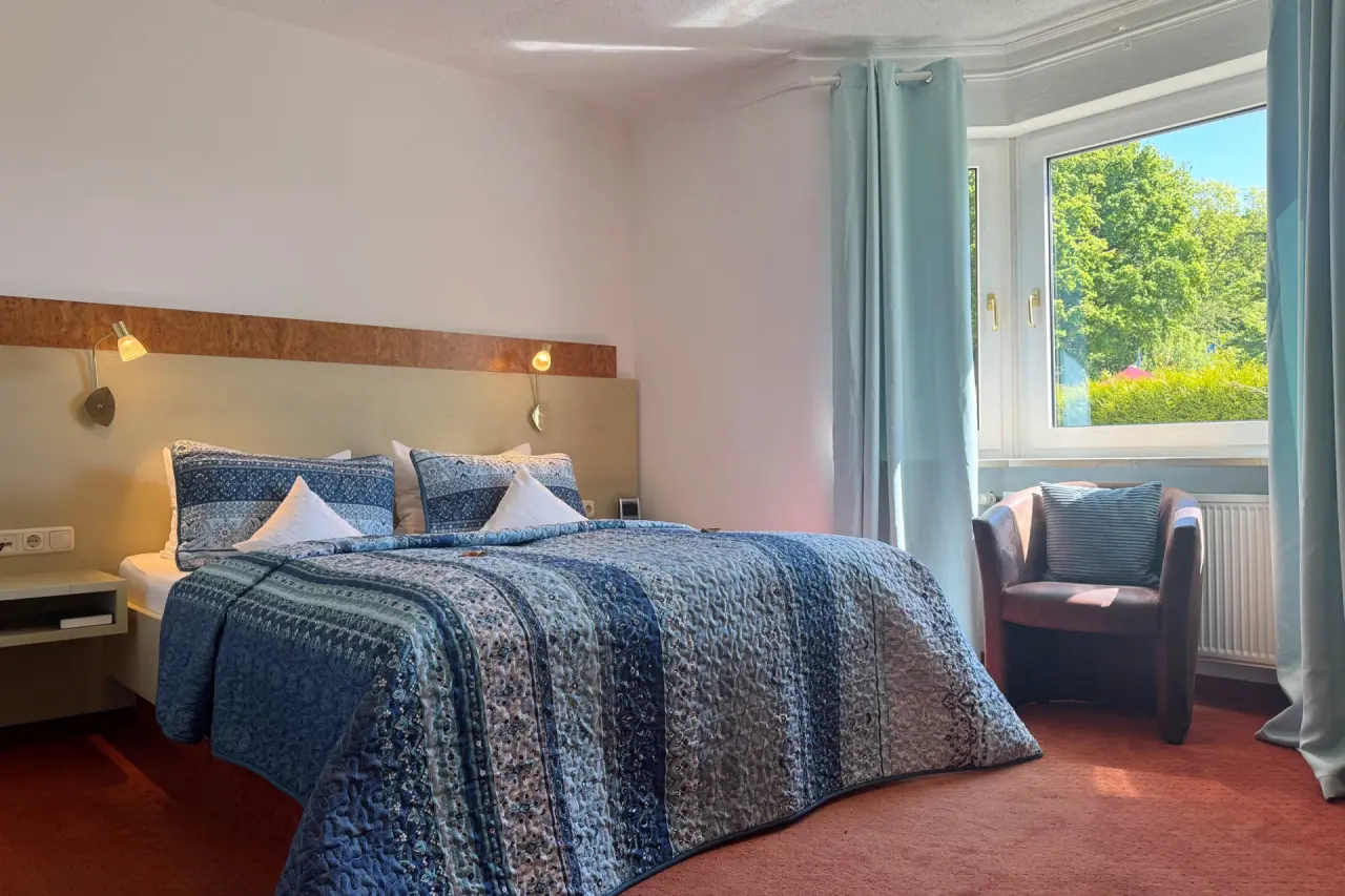 Gemütliches Doppelzimmer im Vitalhotel Sonneck mit blau gemustertem Bett, Sessel am Fenster und Blick ins Grüne.