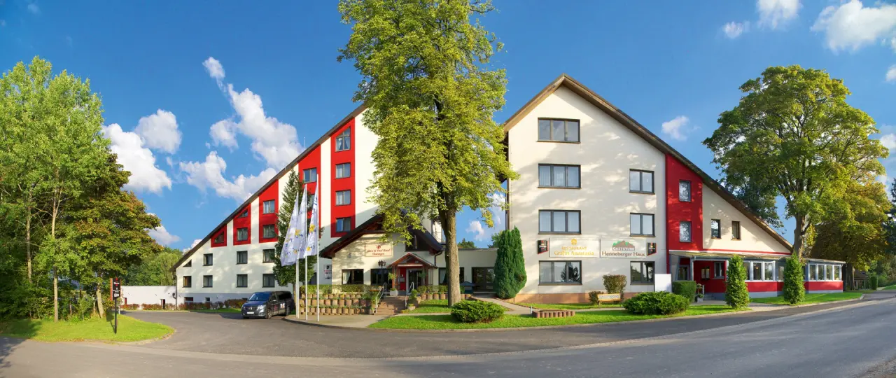 Aussenansicht des AKZENT Aktiv & Vital Hotel Thüringen mit roter und weisser Fassade, umgeben von gruenen Baeumen unter blauem Himmel.