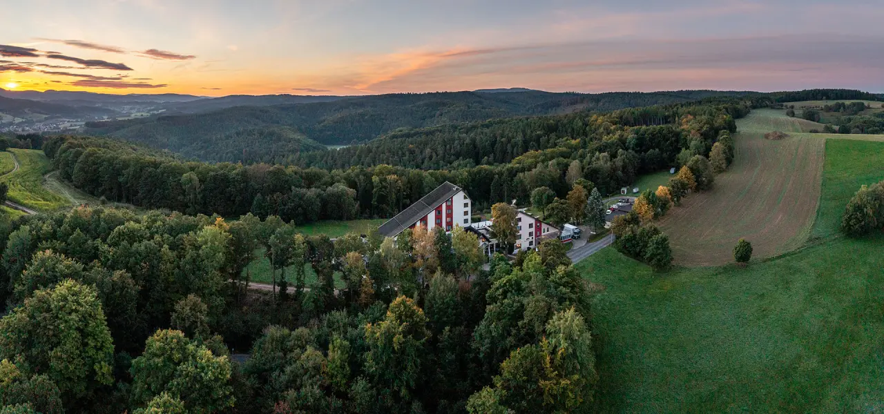 Panoramablick auf das AKZENT Aktiv & Vital Hotel Thüringen inmitten grüner Wälder und Felder bei Sonnenuntergang.