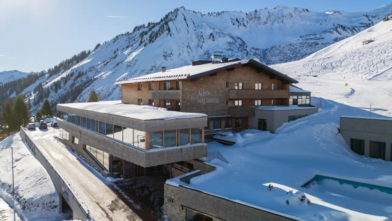 Alpenresort Walsertal im Winter: Hotelgebäude umgeben von Schnee und majestätischen Bergen mit Skipisten.