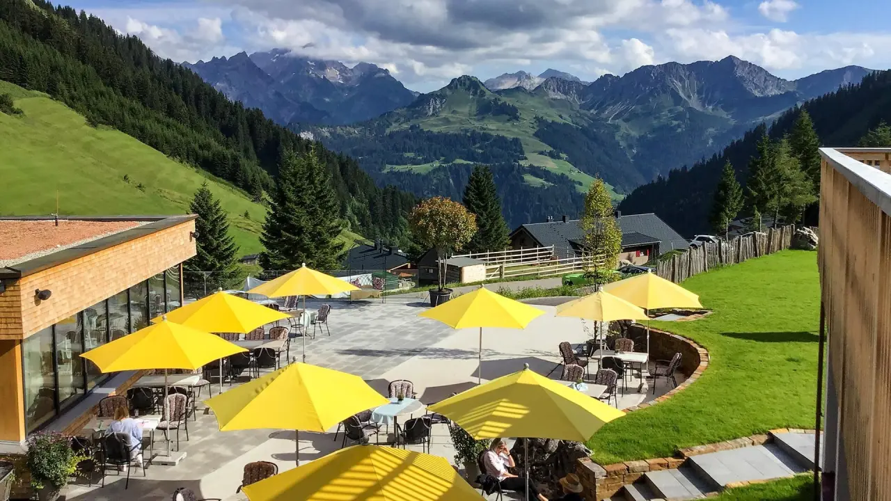 Sonnige Restaurantterrasse mit gelben Sonnenschirmen und atemberaubendem Bergpanorama im Alpenresort Walsertal.