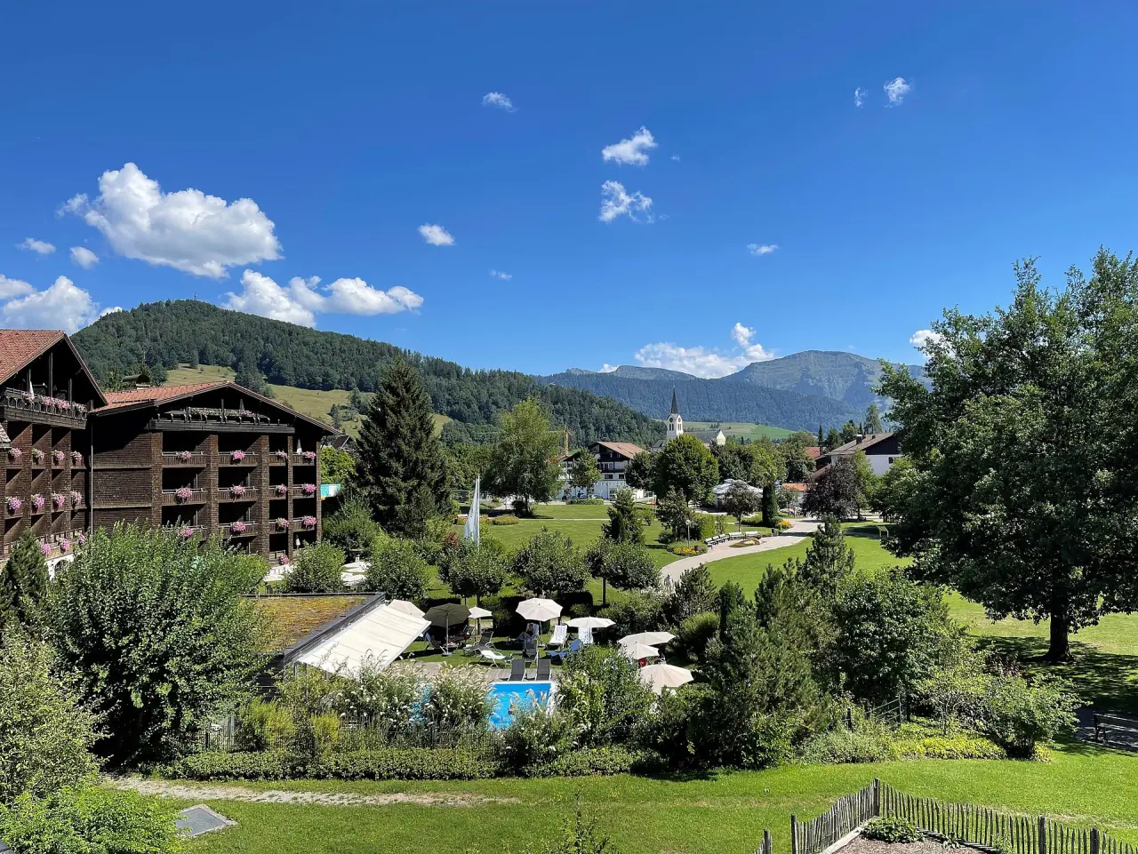 Aussenansicht des Lindner Hotel Oberstaufen Parkhotel mit Garten, Aussenpool und Alpenpanorama unter blauem Himmel.