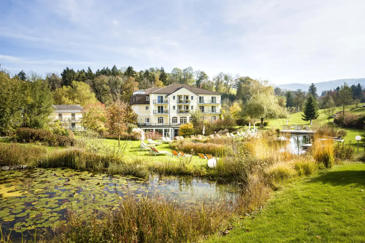 RelaxResort Kothmühle: Idyllisches Hotelgebäude mit Naturteich und Liegewiese, umgeben von herbstlicher Landschaft.