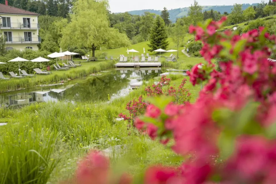Naturbadeteich mit Liegewiese und Hotelgebäude im RelaxResort Kothmühle, umgeben von grüner Landschaft und Blumen.