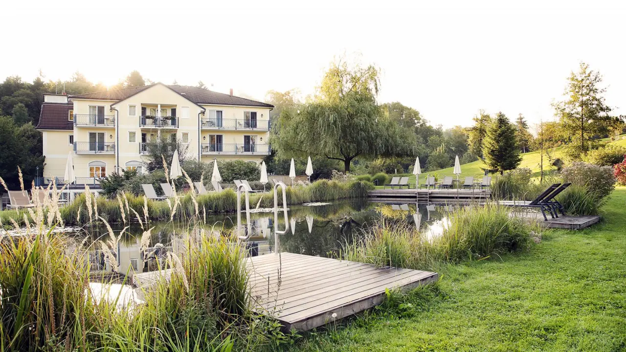 Naturschwimmteich mit Liegewiese und Hotelgebäude des RelaxResort Kothmühle im Abendlicht.