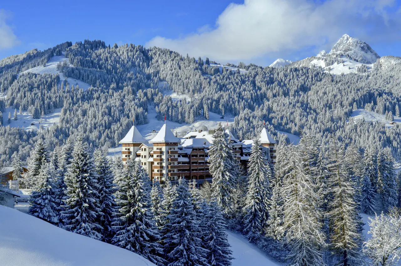 Majestätisches The Alpina Gstaad Hotel im Winter, umgeben von verschneiten Tannen und Schweizer Alpenpanorama.