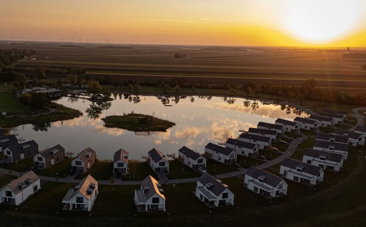 Luftaufnahme des VILA VITA Pannonia Resorts bei Sonnenuntergang mit Bungalows am See und Windrädern im Hintergrund.