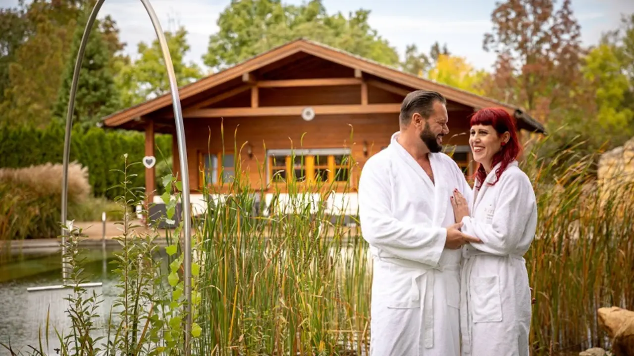 Entspanntes Paar in Bademänteln am Naturbadeteich mit Saunahaus im Wellnessgarten des VILA VITA Pannonia.