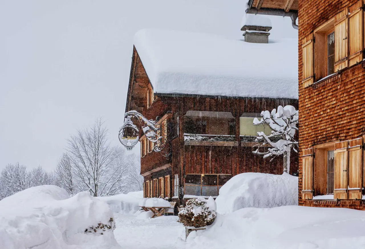 Verschneites Hotel Am Holand im Winter. Traditionelle Holzfassade mit dicken Schneemassen auf dem Dach und im Garten.