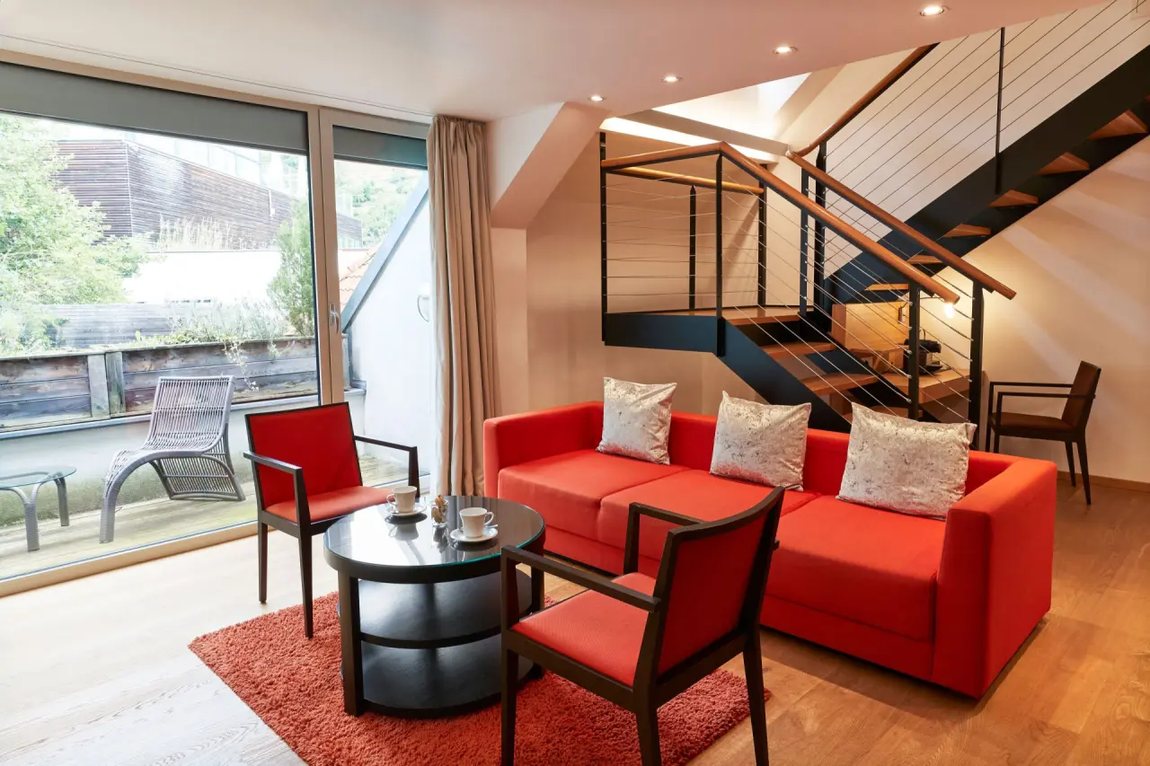 Moderner Wohnbereich einer Suite mit rotem Sofa, Glastisch und Treppe im Steigenberger Hotel and Spa Krems.