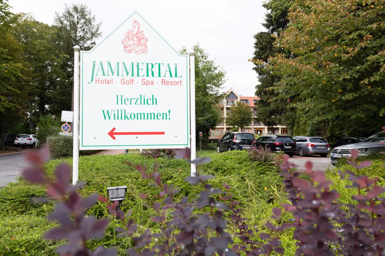 Willkommensschild des Jammertal-Resort mit Hotel, Golf und Spa. Zeigt den Eingangsbereich mit Bäumen und parkenden Autos.
