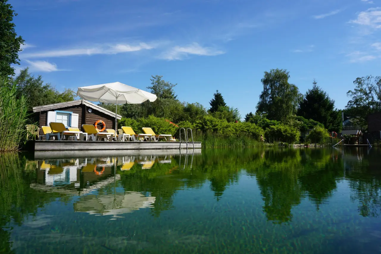 Naturbadeteich mit Holzsteg, Sonnenliegen und Sonnenschirm im Jammertal-Resort unter blauem Himmel.