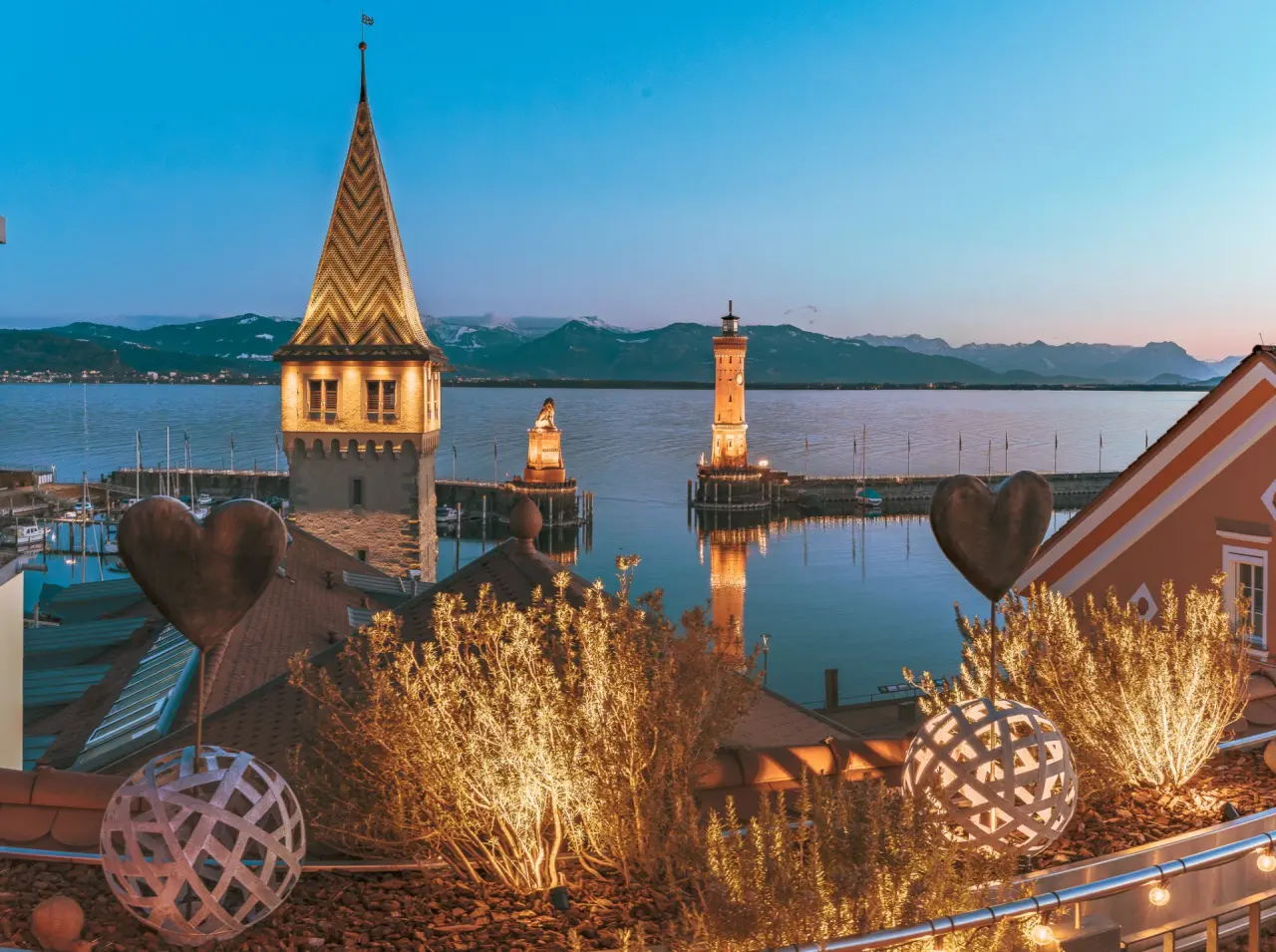 Abendlicher Blick vom Hotel Helvetia auf den Lindauer Hafen mit Leuchtturm, Mangturm und Bodensee vor Alpenpanorama.