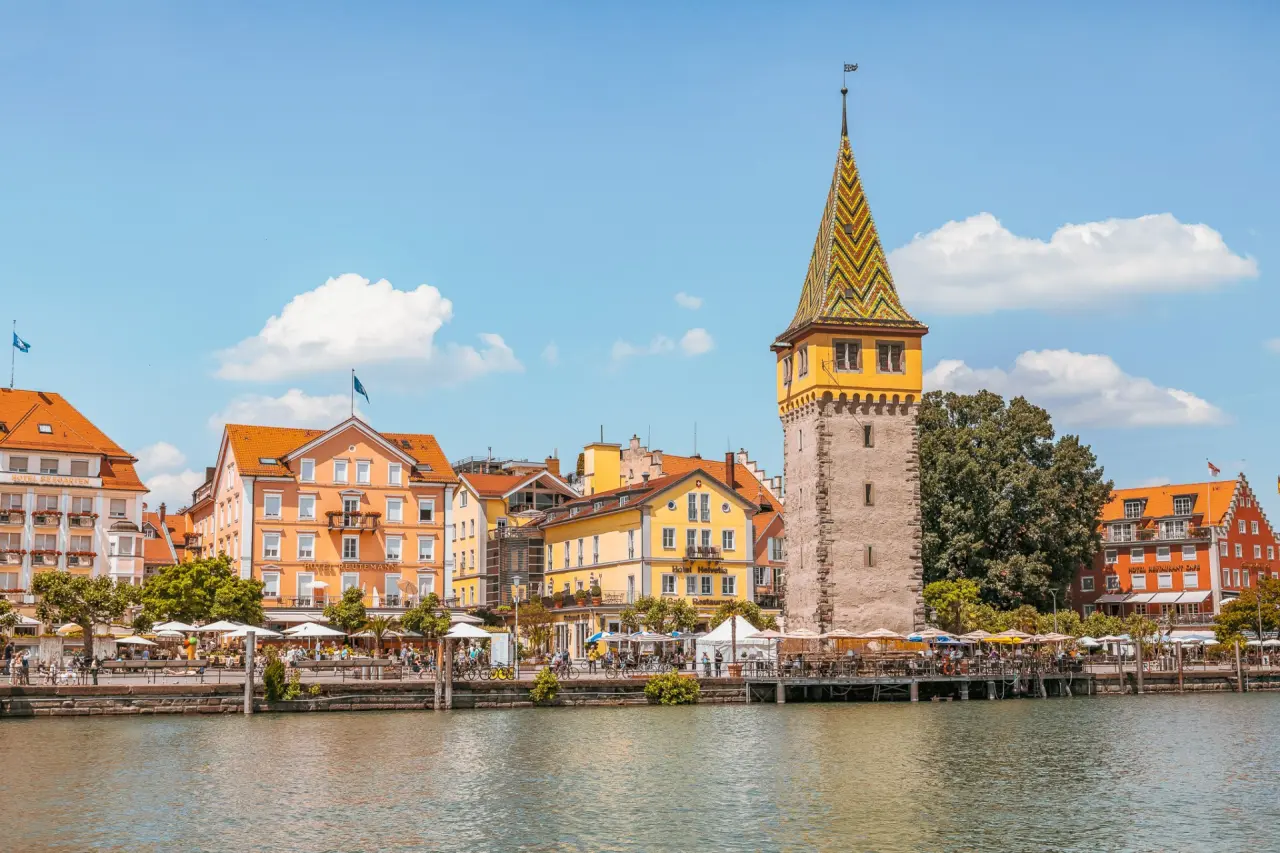 Aussenansicht des Hotel Helvetia am Hafen von Lindau mit dem Mangturm und belebten Terrassen unter blauem Himmel.