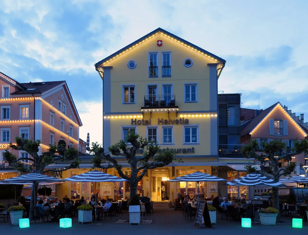 Beleuchtete Aussenansicht des Hotel Helvetia bei Abenddaemmerung mit beleuchteter Restaurantterrasse und Gaesten.