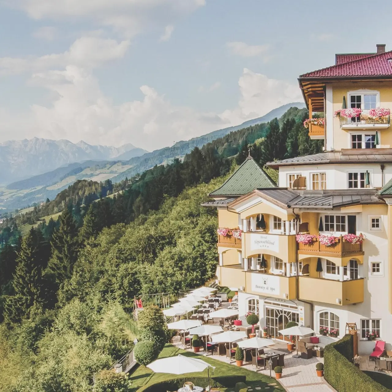 Aussenansicht des Hotel AlpenSchlössl mit blühenden Balkonen und Terrasse vor malerischer Berglandschaft.