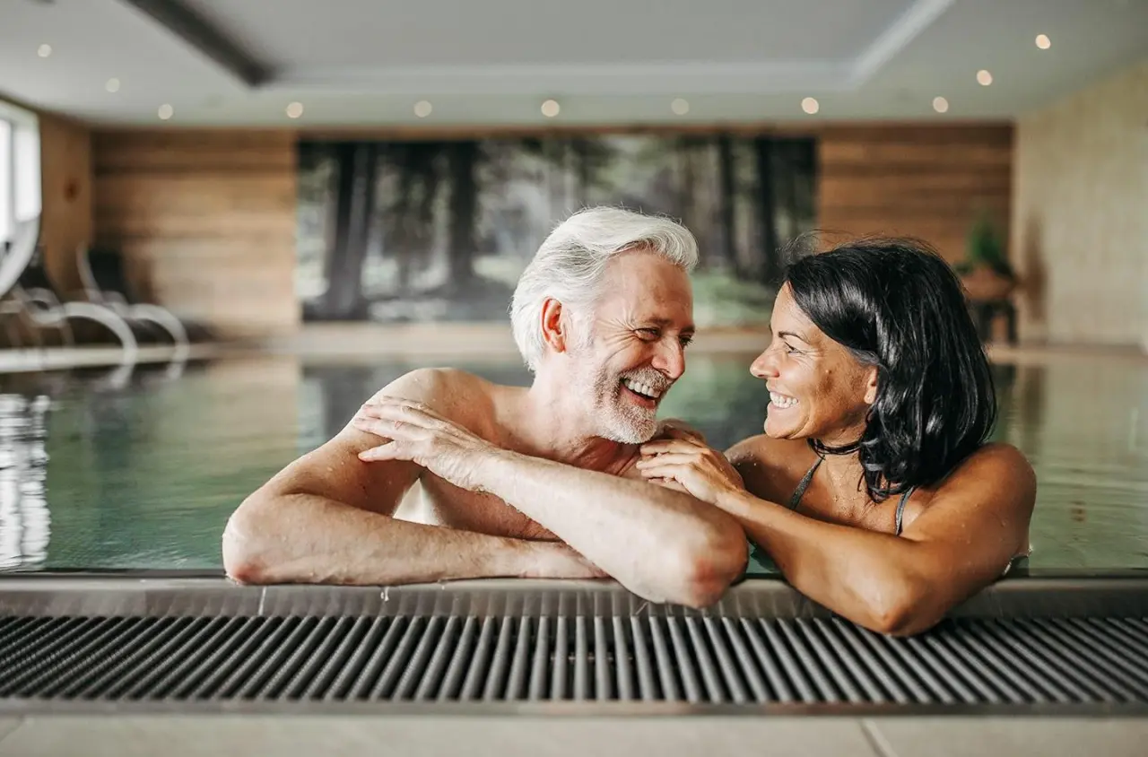 Glückliches Paar entspannt im Innenpool des Hotel Waldfrieden mit Waldmotiv an der Wand.