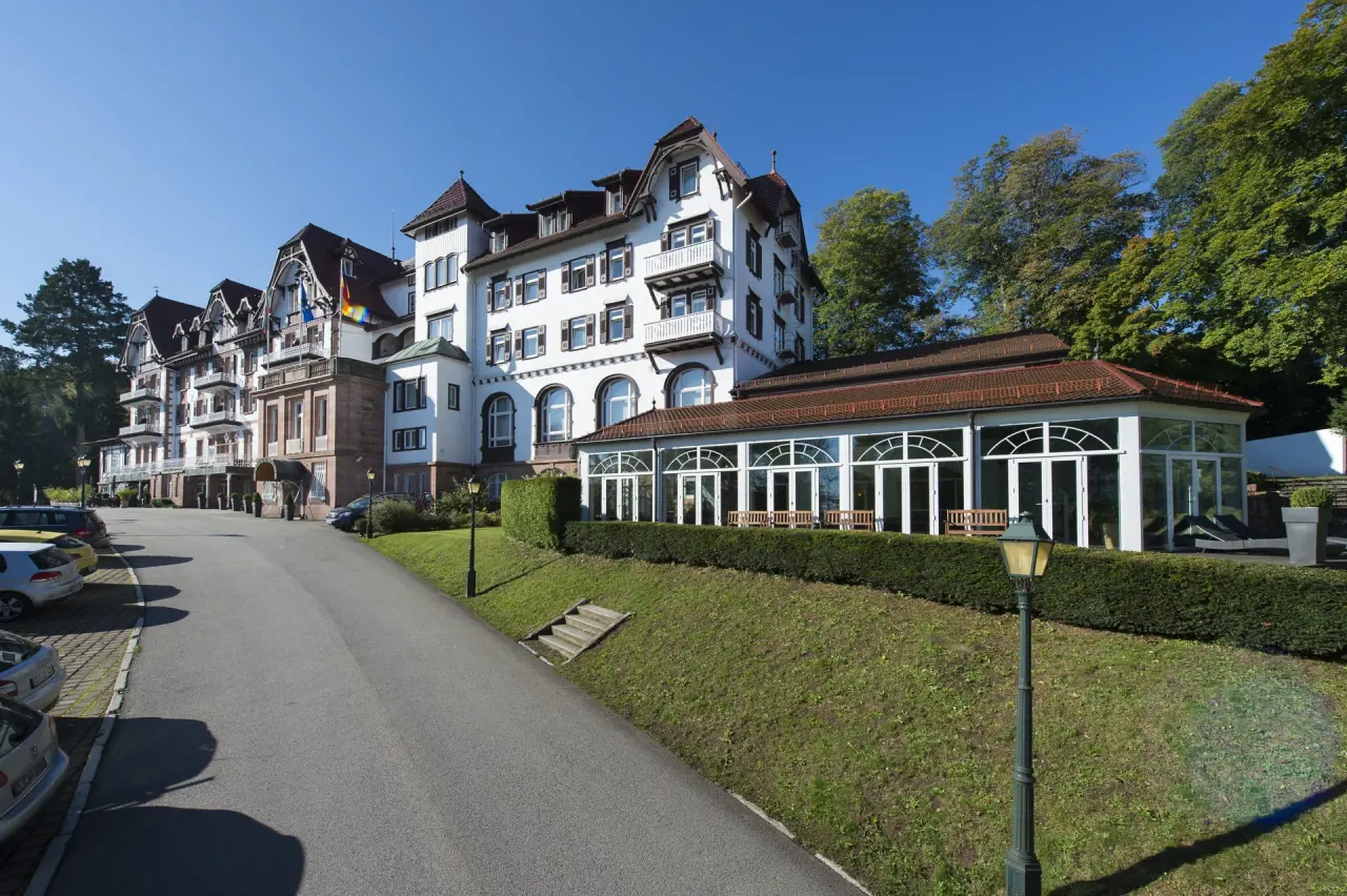 Das historische Wellnesshotel Palmenwald mit seiner eleganten Fassade und dem Wintergarten unter blauem Himmel.