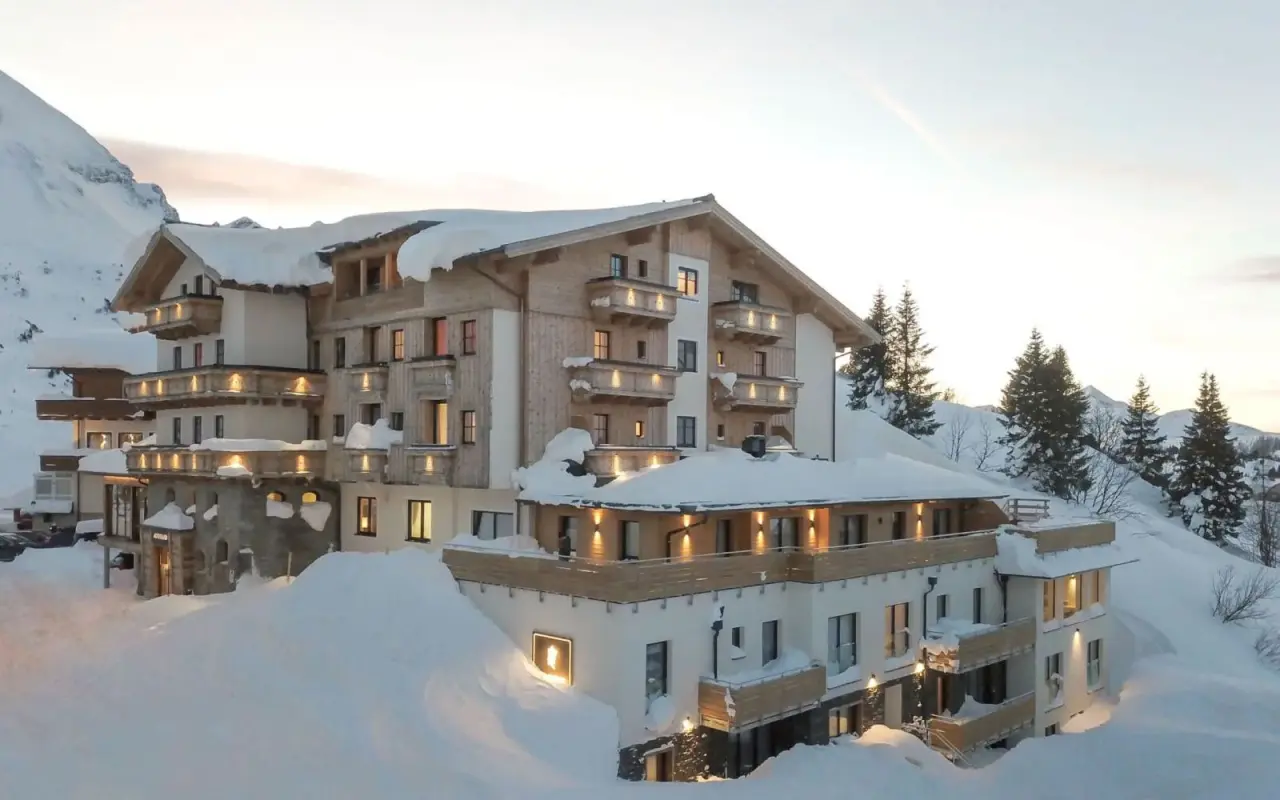 Hotel Alpenland im Winter, umgeben von tiefem Schnee und Bergen bei Sonnenuntergang. Gemütliche Beleuchtung der Balkone und Fenster.