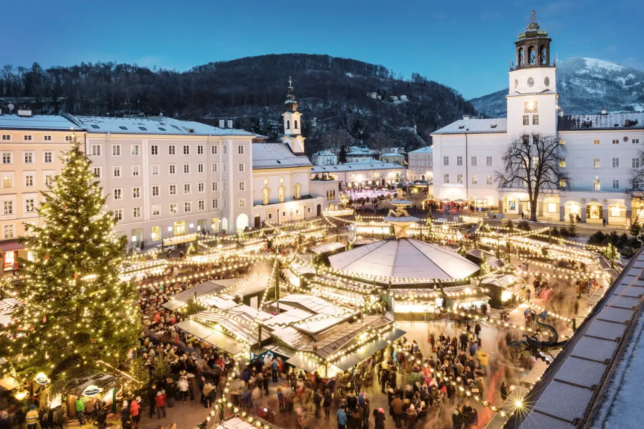 Festlicher Weihnachtsmarkt in Salzburg bei Abenddämmerung, umgeben von historischen Gebäuden und verschneiten Bergen.
